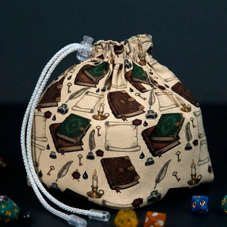 Books and Scrolls dice bag standing on a dark table, cream fabric with leather grimoires, scrolls, candles and quills
