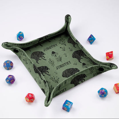 Jimothy sage botanical dice tray at an angle, black raccoons and wildflowers on sage green fabric, colorful dice around it