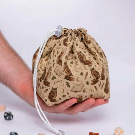 Owl Atheneum dice bag resting in an open palm, tan fabric printed with owls perched on book stacks, moons and reading glasses