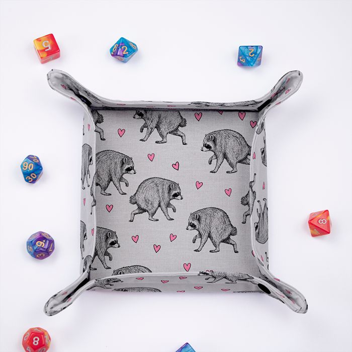 Jimothy raccoon dice tray from above, sketched raccoons and pink hearts on light grey fabric, colorful dice around it