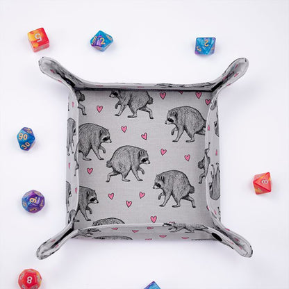 Jimothy raccoon dice tray from above, sketched raccoons and pink hearts on light grey fabric, colorful dice around it