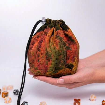 Autumn Forest dice bag held in a hand, painterly print of orange and green fall trees, black drawstring with cord lock