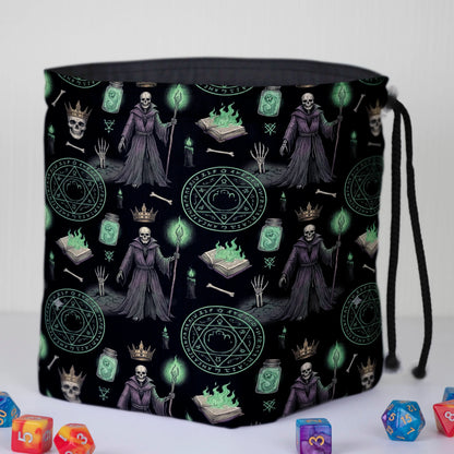 Lich dice bag standing open on the table — robed skeleton liches, spellbooks with green flame and summoning circles, polyhedral dice around