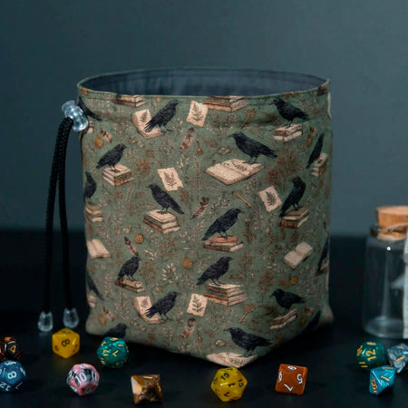 Ravensward dice bag standing open on its base, sage green fabric with black ravens perched on old books, grey lining inside