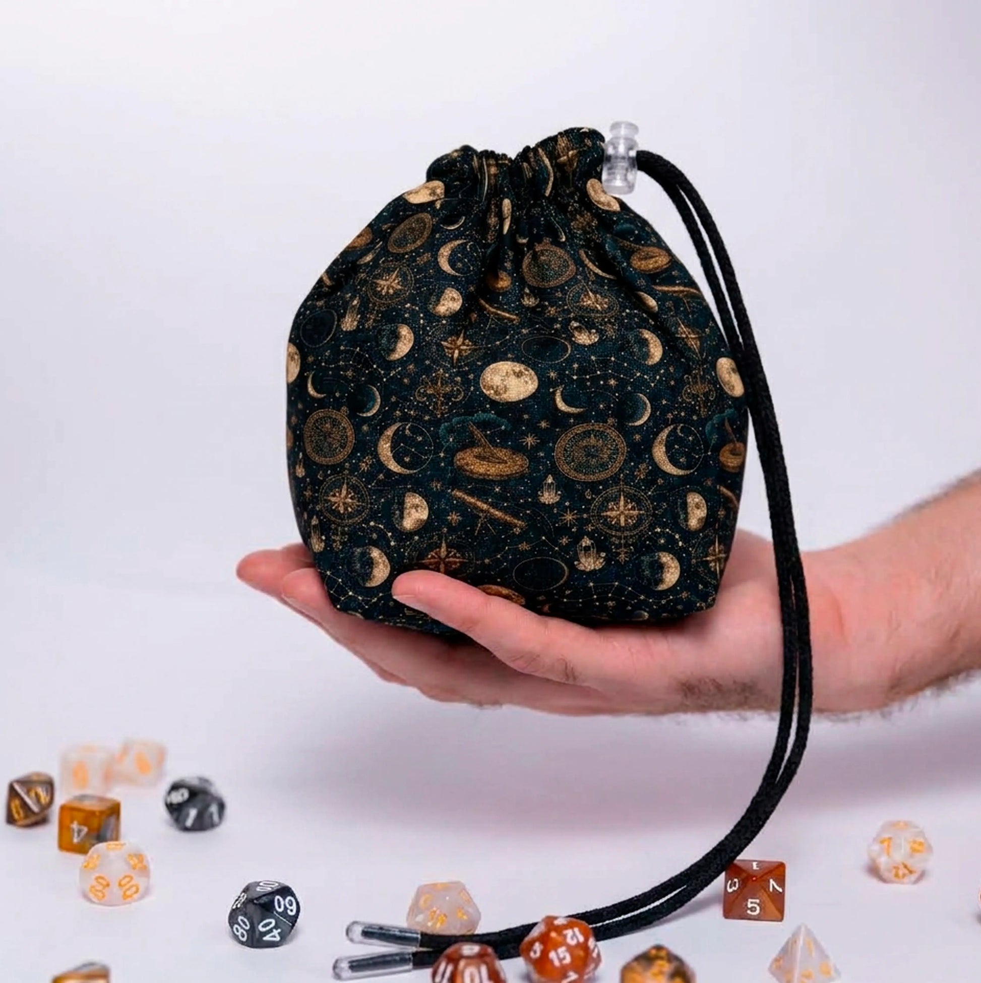Celestial Almanac dice bag held in one hand, dark teal fabric with gold moon phases, constellations and sundials