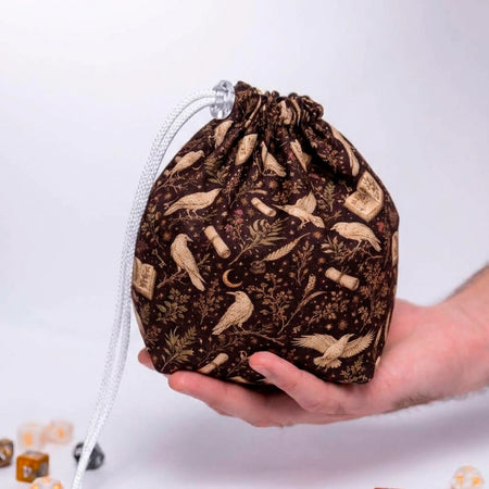 Raven's Codex dice bag resting in a palm, dark brown fabric with cream ravens, open books, scrolls and a crescent moon