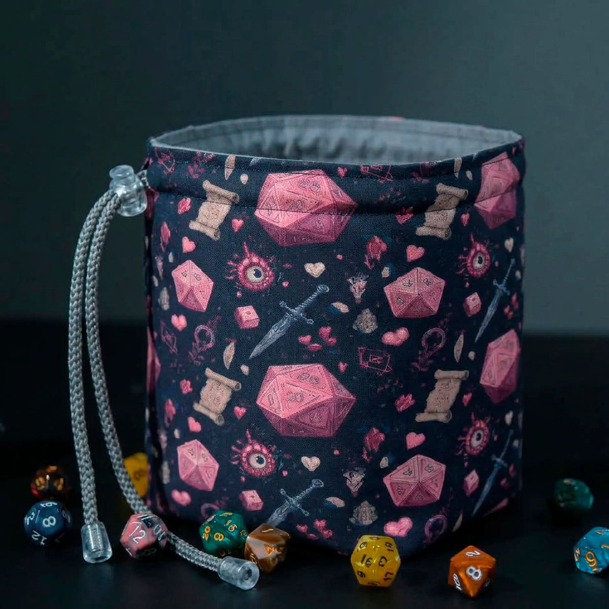 Crimson D20 dice bag standing open on its square base, navy fabric with pink d20 and dagger print, grey cotton lining, dice scattered around