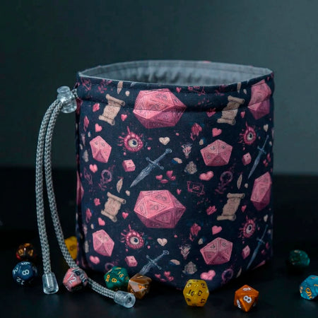 Crimson D20 dice bag standing open on its square base, navy fabric with pink d20 and dagger print, grey cotton lining, dice scattered around