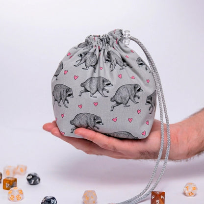 Jimothy raccoon dice bag in light grey with tiny pink hearts print, held in a hand with DnD dice