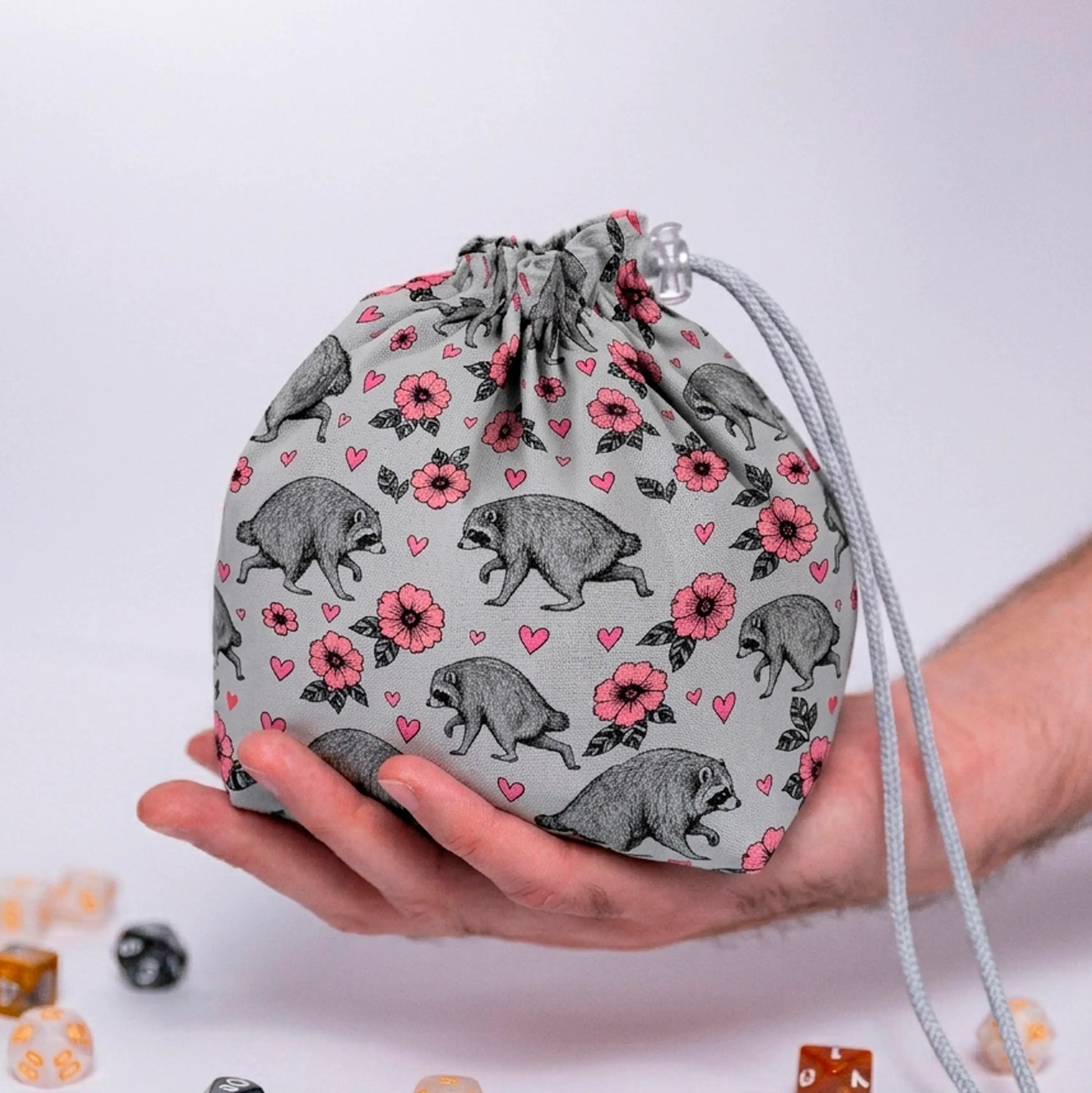 Jimothy raccoon dice bag in grey with pink blossoms and hearts print, held in a hand with DnD dice