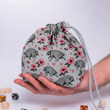 Jimothy raccoon dice bag in grey with pink blossoms and hearts print, held in a hand with DnD dice