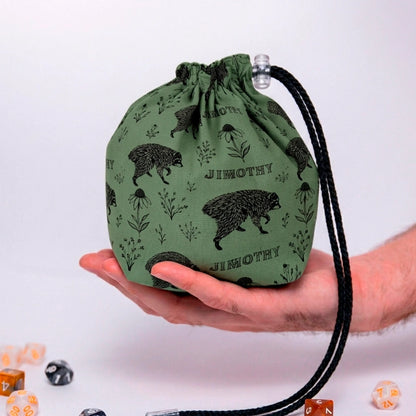 Jimothy raccoon dice bag in sage green botanical print with JIMOTHY lettering and wildflowers, held in a hand with DnD dice