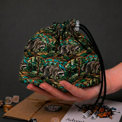 Jimothy raccoon dice bag in dark stained glass forest print with greens, teal and amber, held in a hand