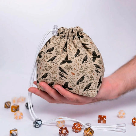 A Murder of Crows dice bag held in one hand, cream fabric printed with black crows and autumn branches, dice on the table below
